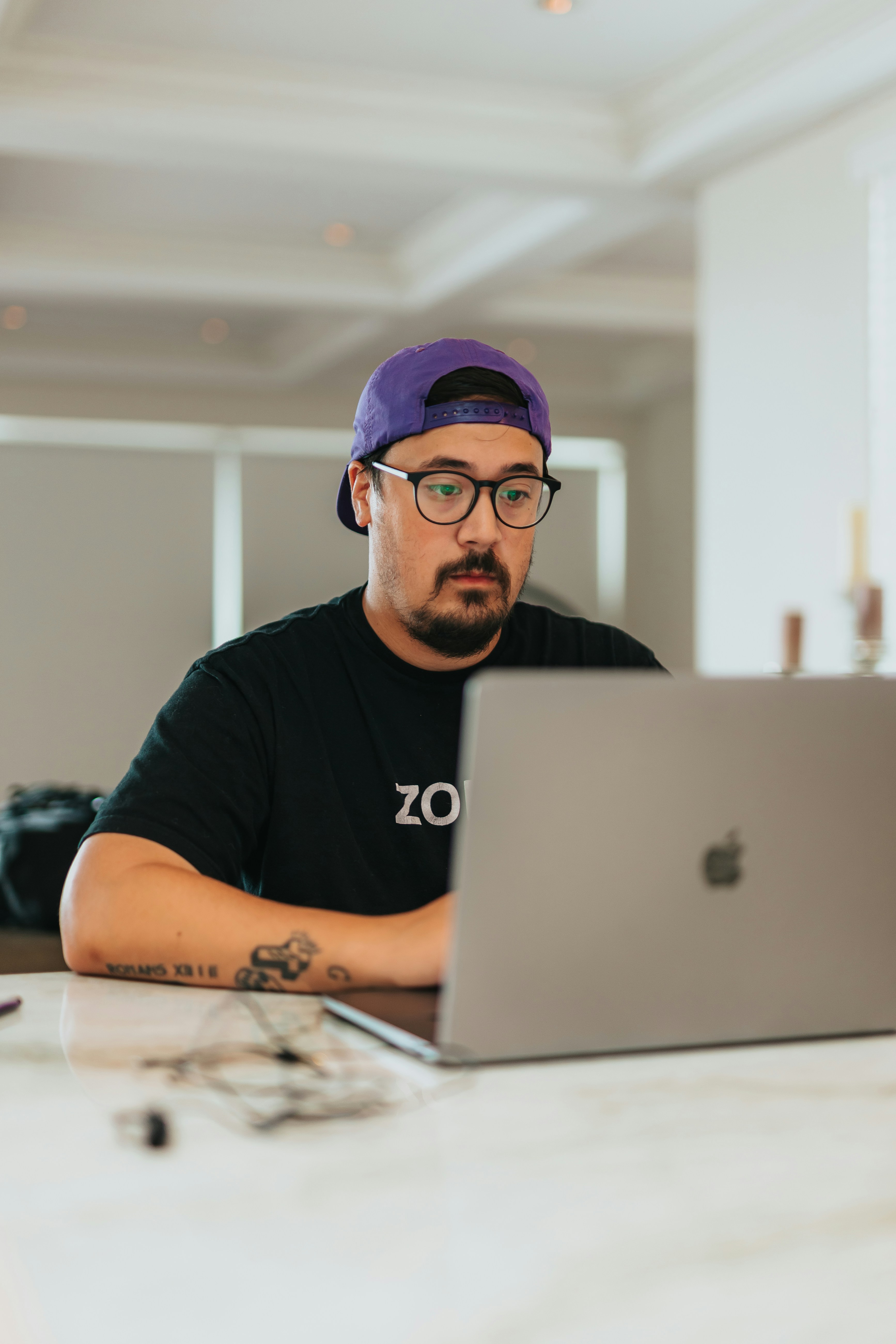 A person wearing a purple cap, a black t-shirt and glasses is sitting and working at a laptop.